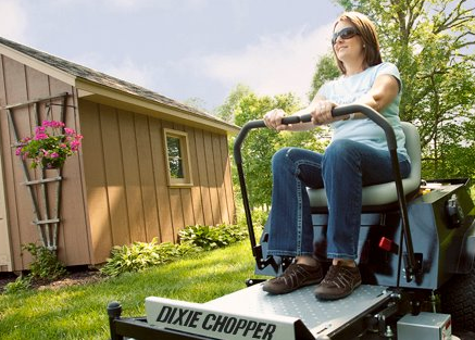 Woman driving a Dixie Chopper lawnmower on a green lawn near a brown shed with flowers.