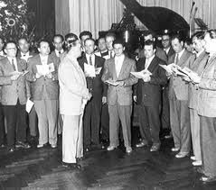 Group of men in suits standing, some holding papers, near a piano in a room.