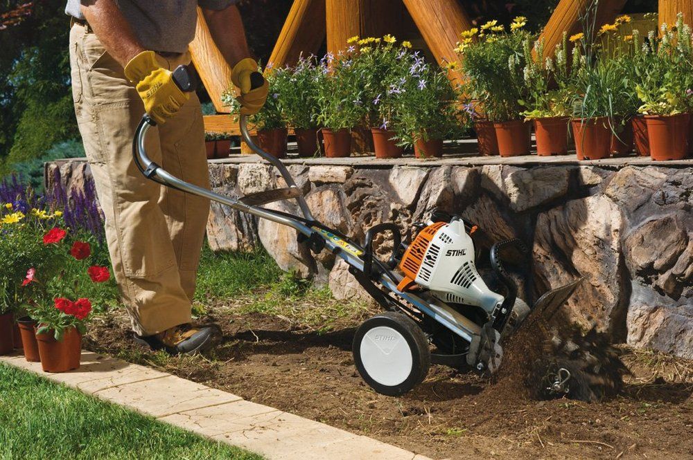 A person using a tiller to cultivate soil next to a stone wall and potted flowers.