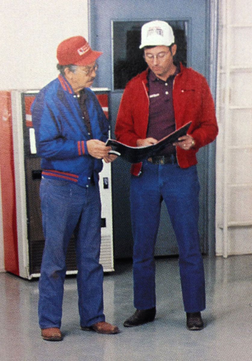 Two men in casual wear reviewing a document in a facility. One in red jacket, the other in a blue jacket.