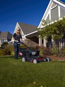 Woman mowing a lawn with a black and red lawnmower in front of a house on a sunny day.