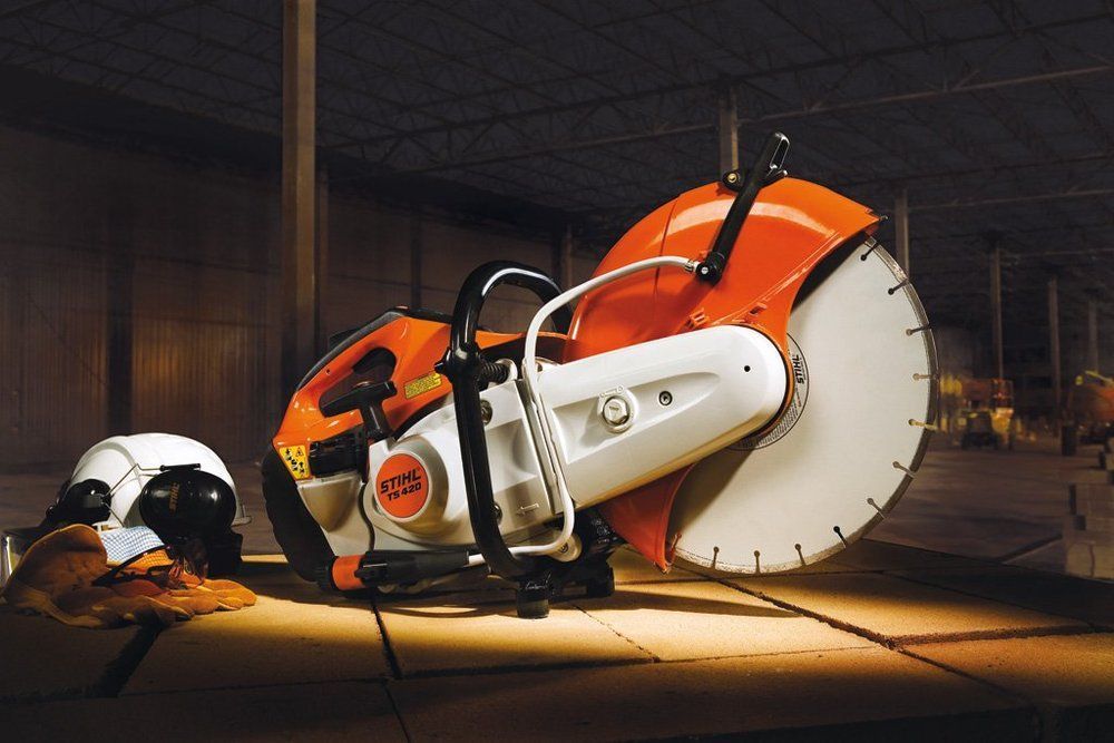Orange and white power cut-off saw with large blade. A hard hat and gloves sit nearby, on a concrete surface.