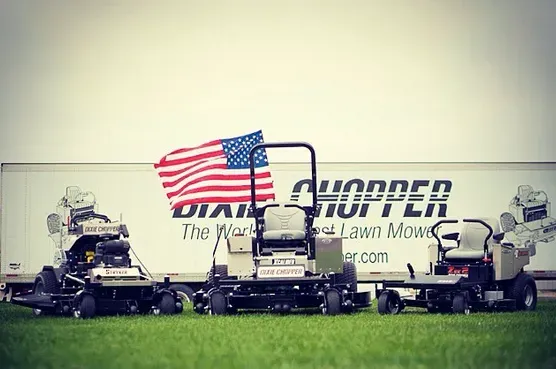 Three Dixie Chopper lawn mowers parked on grass in front of a trailer with an American flag.