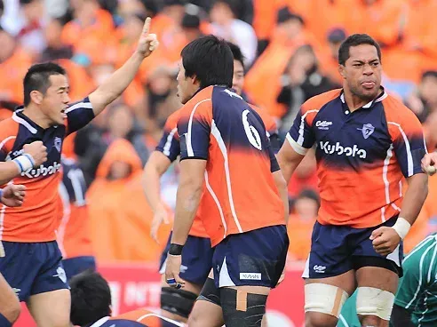 Rugby players in orange and navy uniforms celebrate on a field; one raises a fist.