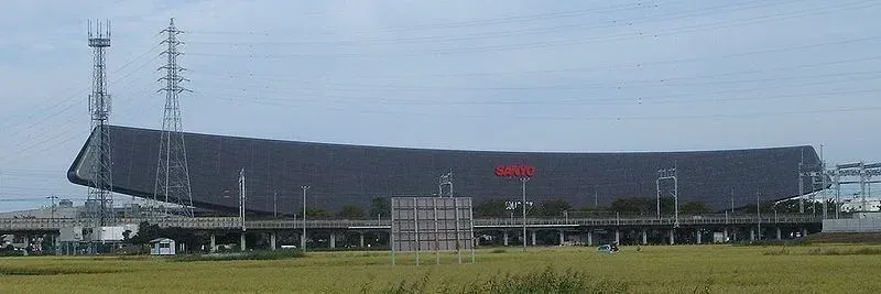 A large, dark-colored stadium with antennas in a grassy field under a pale blue sky.