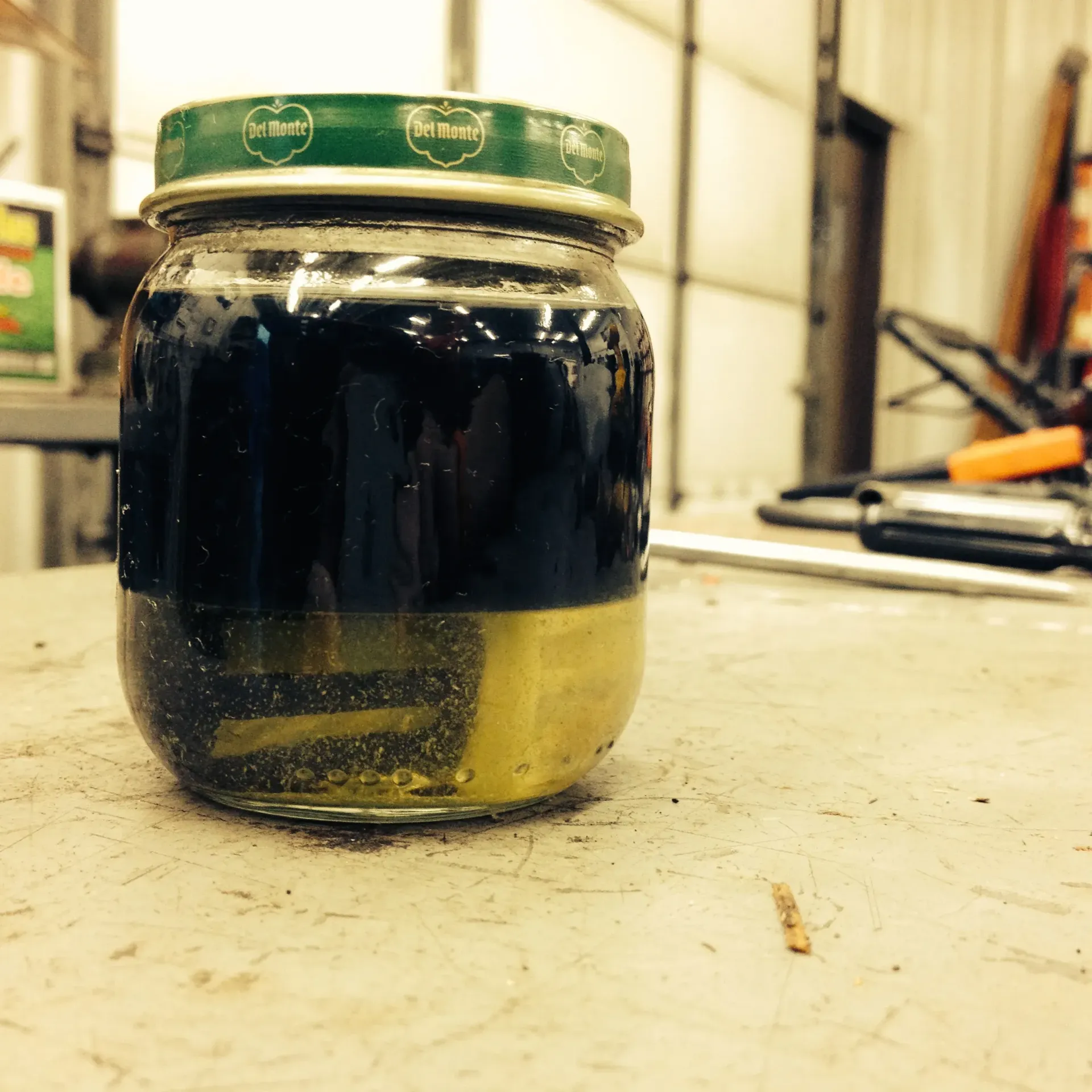 Jar of dark liquid with a lighter colored layer settled at the bottom, on a cluttered work surface.