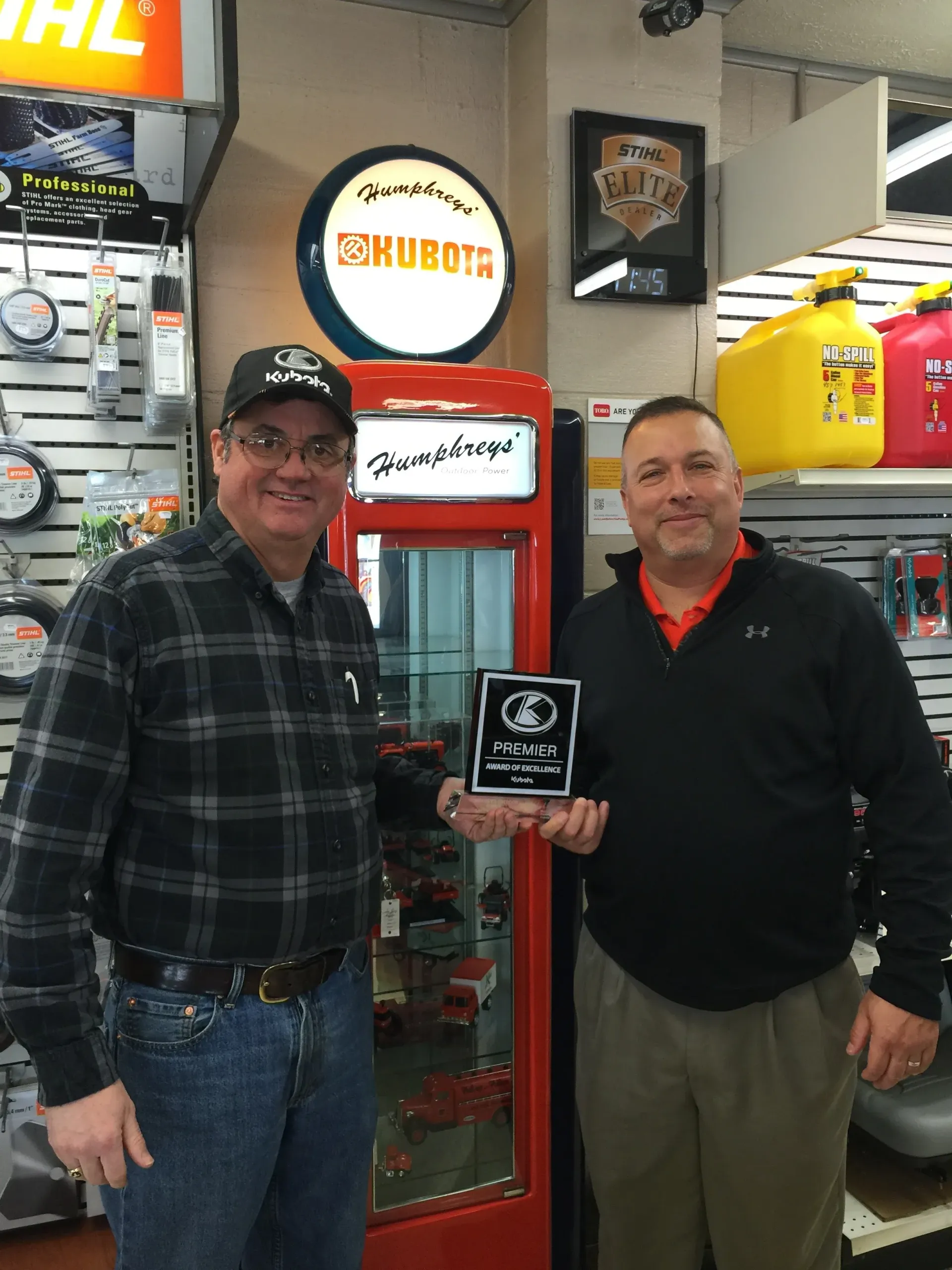 Two men in a store holding an award, near a Kubota sign and display case.