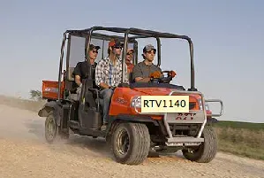 Four people ride in an orange utility vehicle on a dirt road.