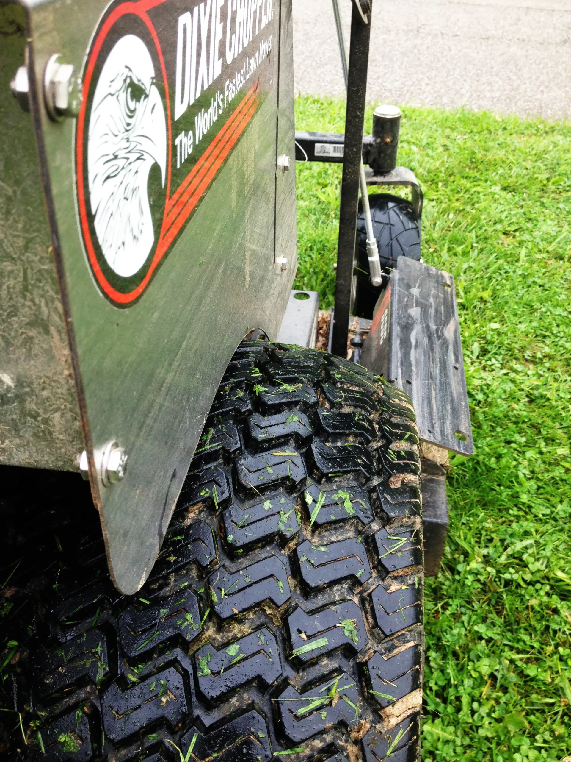 Side view of a mower with a black tire on green grass, next to a silver metal panel with a logo.