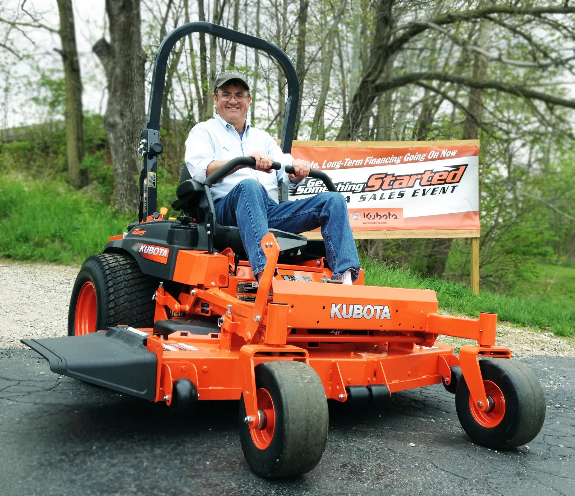 Man seated on an orange Kubota zero-turn mower in front of a sign.