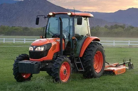 Orange Kubota tractor with a rotary cutter on a grassy field with mountains in the background.