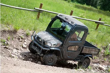 Gray utility vehicle driving up a dirt hill, near a wooden fence.