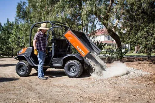 Man in hat unloading gravel from a Kubota utility vehicle with an orange bed in a yard.