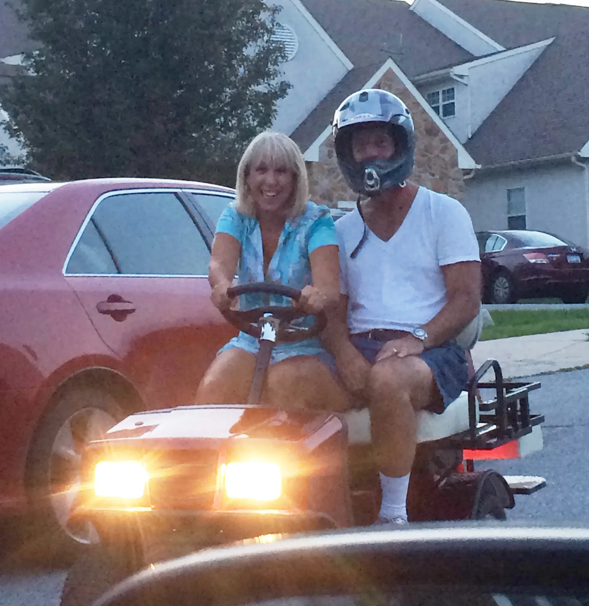 Woman and man on a lawn mower, smiling. Man in helmet, woman at wheel. Red car and houses in background.