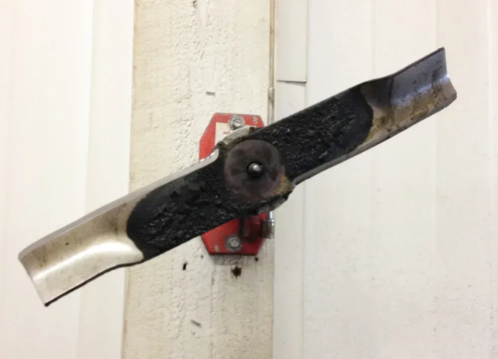 A dirty, damaged lawnmower blade mounted on a white column with a red bracket.