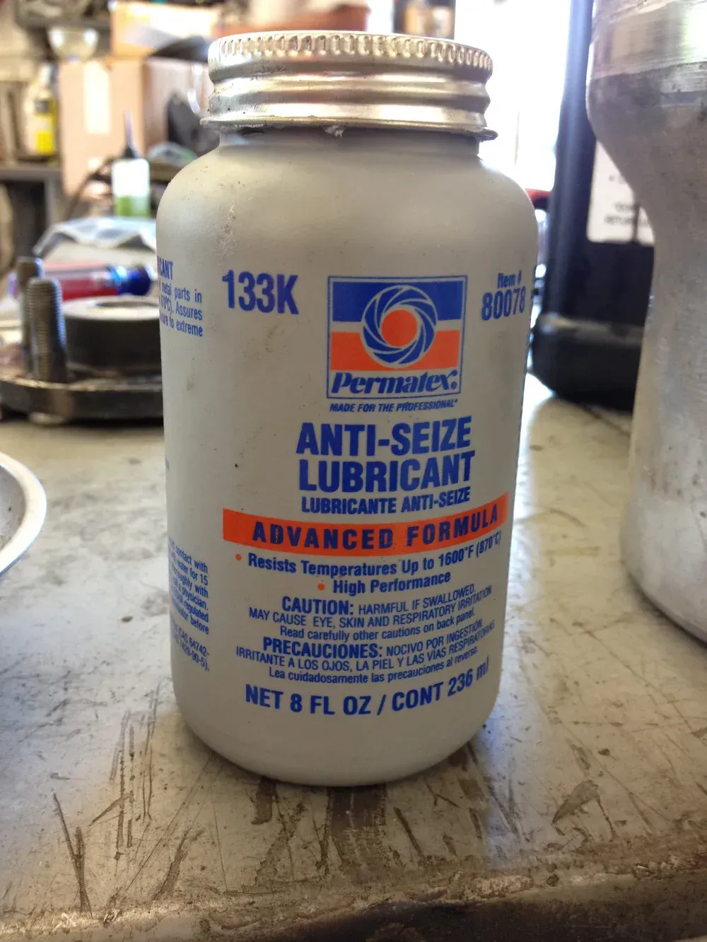 A jar of Permatex Anti-Seize Lubricant on a cluttered workbench. Blue and orange label, silver cap.