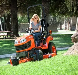 Woman driving an orange tractor, mowing grass in a grassy area with trees and picnic tables.