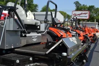 Lawn mowers displayed outside a store; some are orange, others are silver with black seats, under a sign.