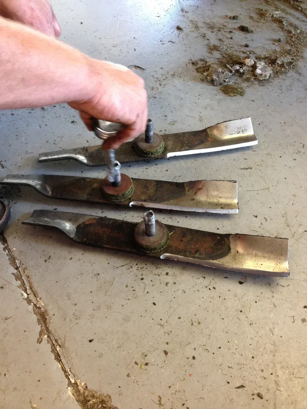 Person's hand pointing at three lawnmower blades, covered in grass, on a concrete surface.