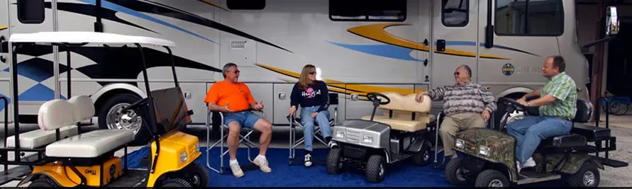 Four people sitting near golf carts and a motorhome. Two sit on chairs, the others in carts.