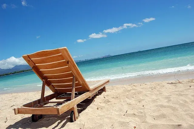 Wooden beach chair on a sandy beach, overlooking turquoise ocean under a bright blue sky.
