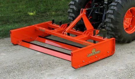 Orange tractor box blade attached to a tractor, on gravel.