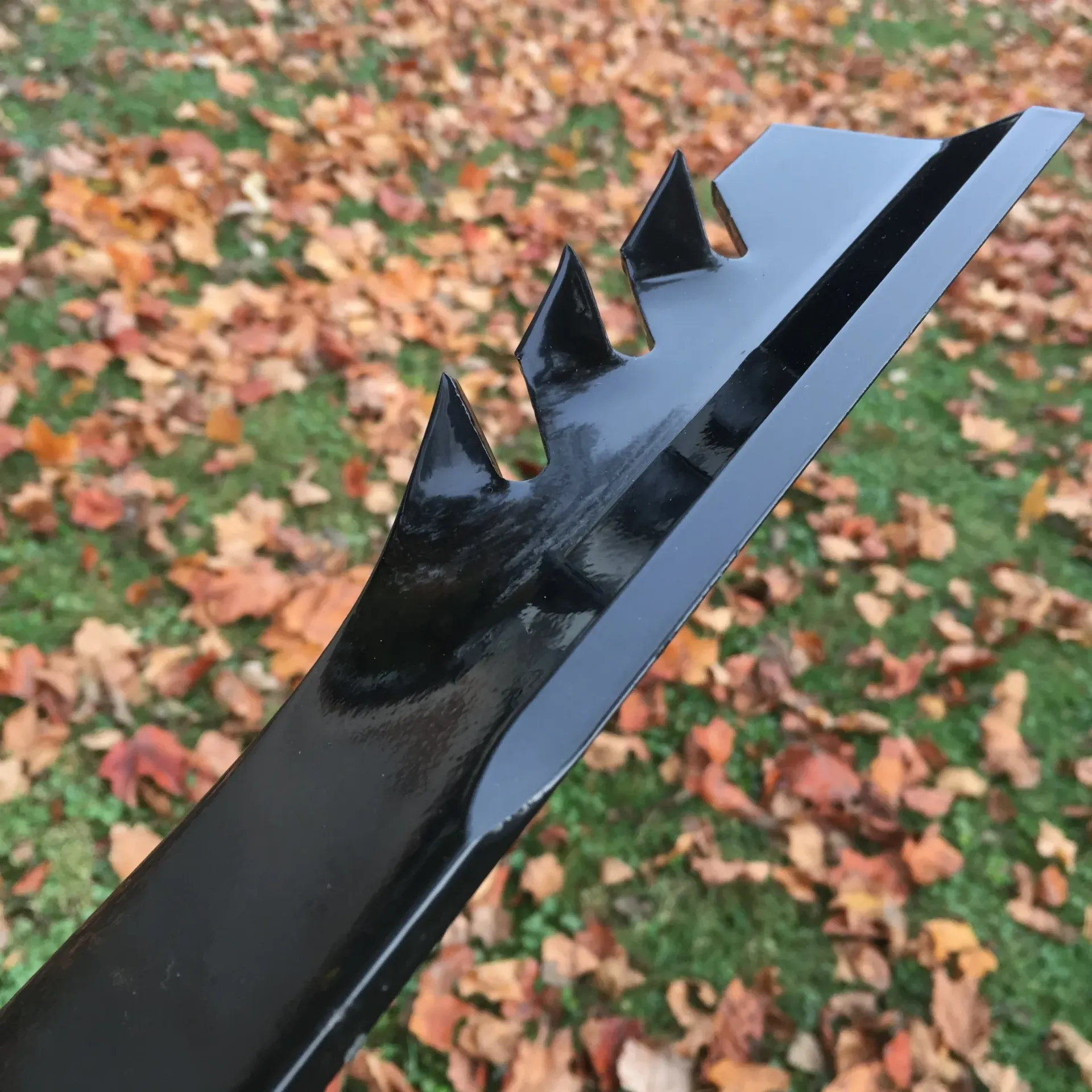 Black lawn mower blade with angled, serrated edge, on fallen leaves.