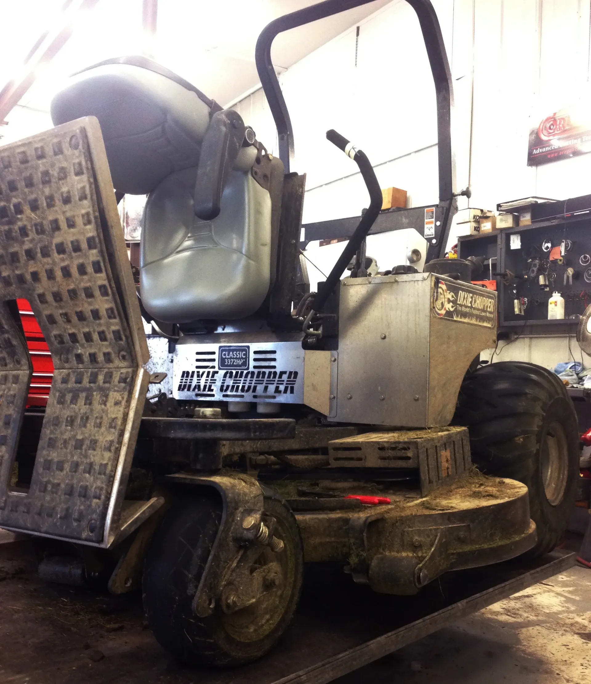 Gray commercial zero-turn lawn mower in a garage, with a diamond-plate access ramp and large tires.