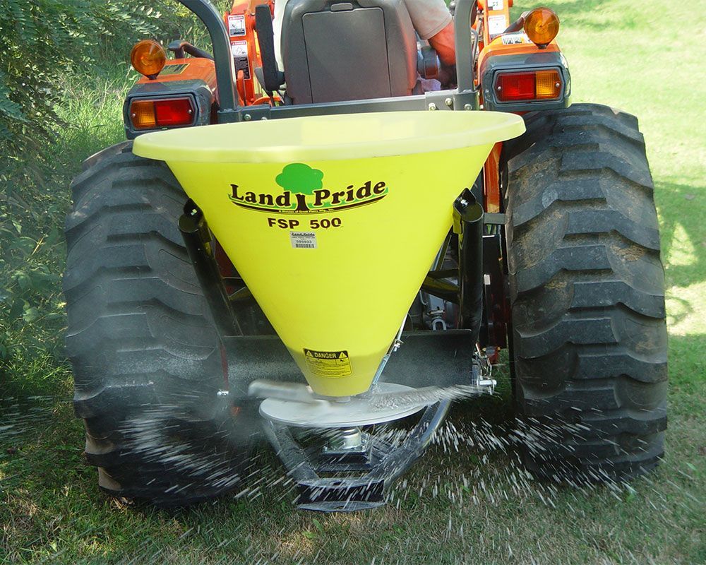 Yellow Land Pride spreader attached to orange tractor, spreading material onto grass.