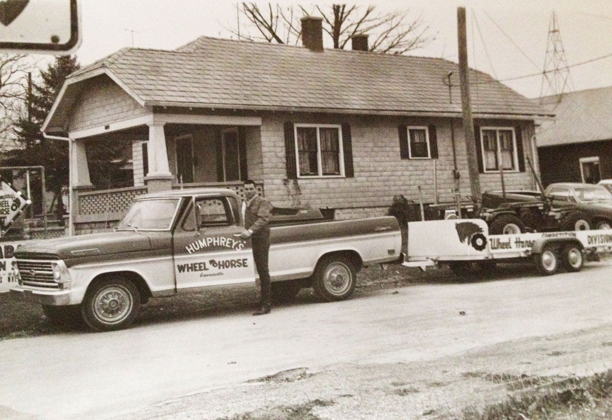 Vintage pickup truck with trailer carrying a car in front of a house.