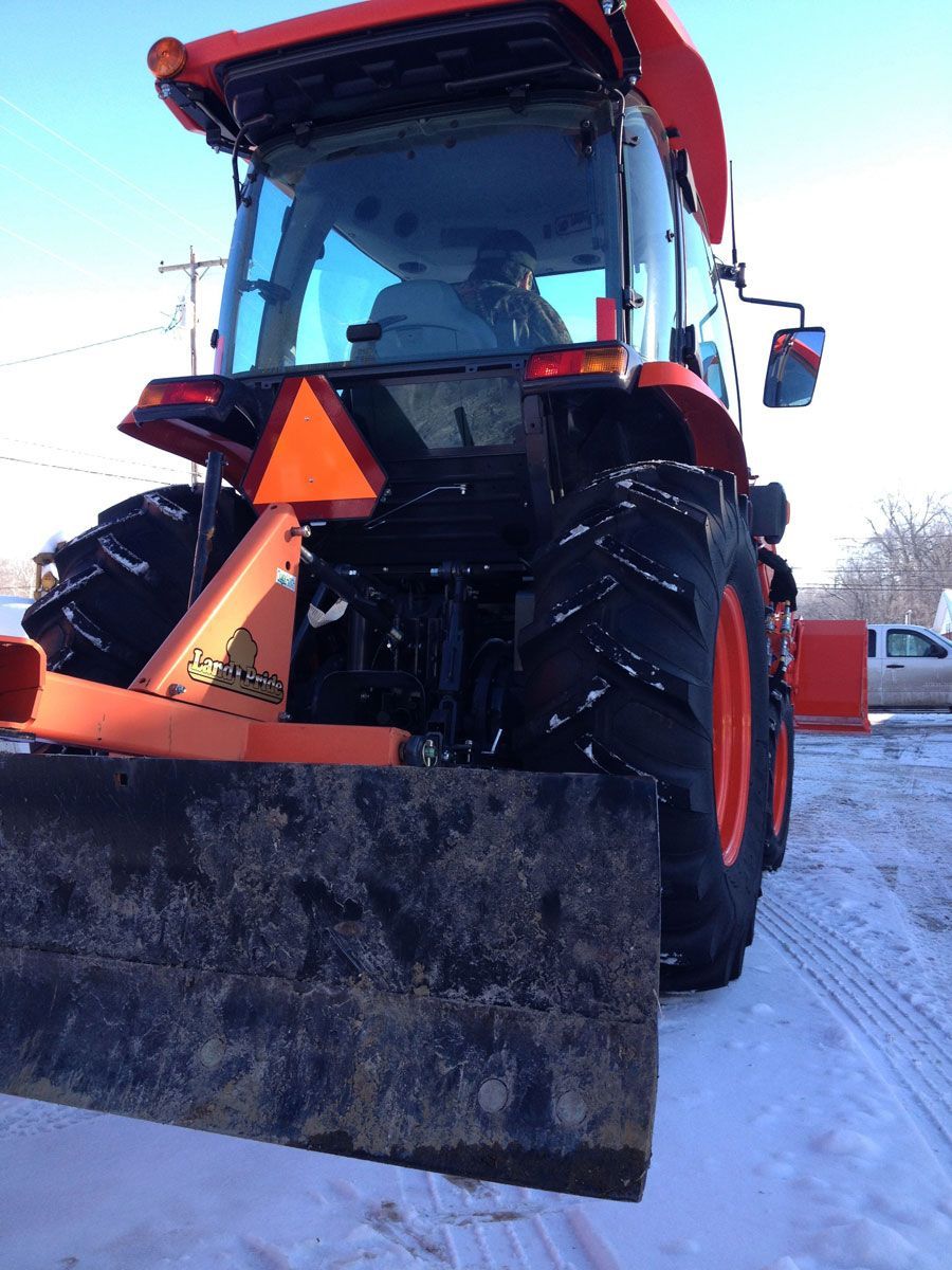 Orange tractor with blade, rear view, on snow-covered ground.