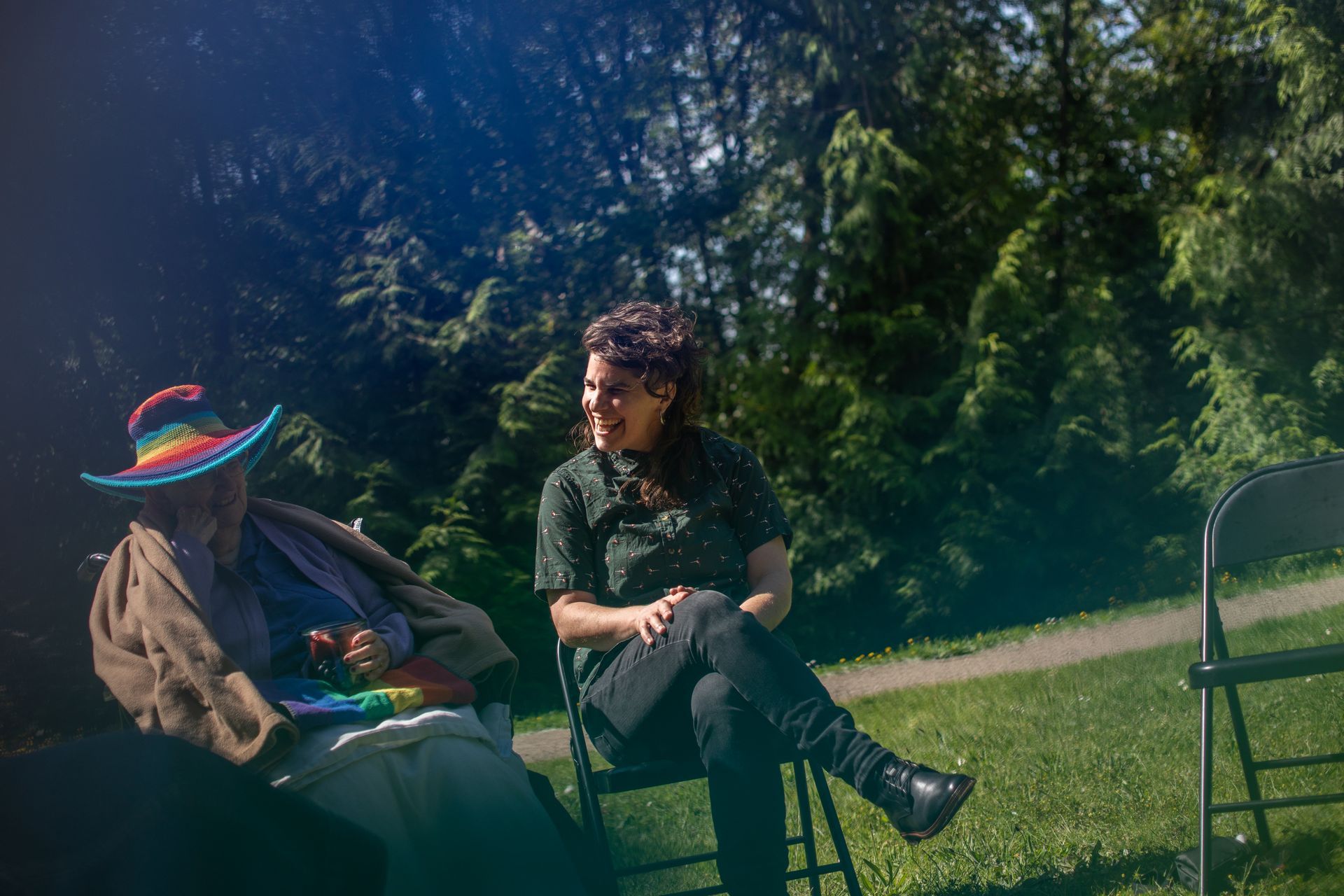 Two persons are sitting in chairs in a park . One of the person is wearing a rainbow hat.