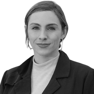 Woman with fair skin wearing a black blazer and hoop earrings, looking at the viewer.