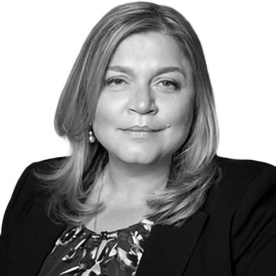 Woman with shoulder-length blonde hair, wearing a dark blazer and patterned top, looking forward.