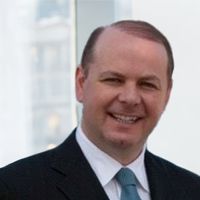 Man in a suit smiles. Blue tie, white shirt, and a window in the background.