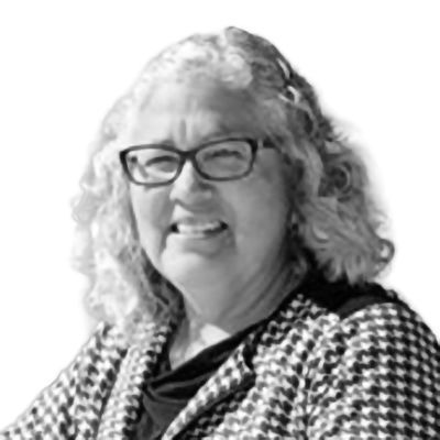 Woman with curly gray hair, wearing glasses and patterned jacket, smiling.