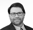 Professional headshot of a person wearing glasses, a suit, and a tie against a plain white background.