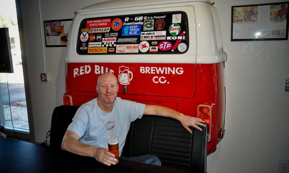 A man is sitting in front of a red bus that says brewing co.