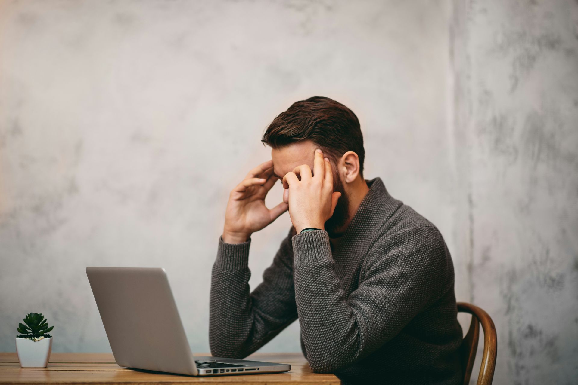 Stressed man sitting at a desk with laptop, holding his head in his hands