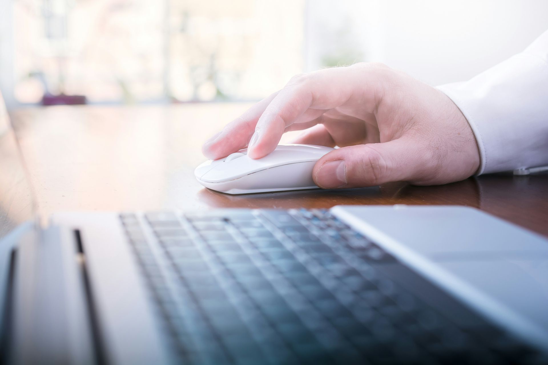 Hand using a white mouse beside a laptop keyboard on a desk