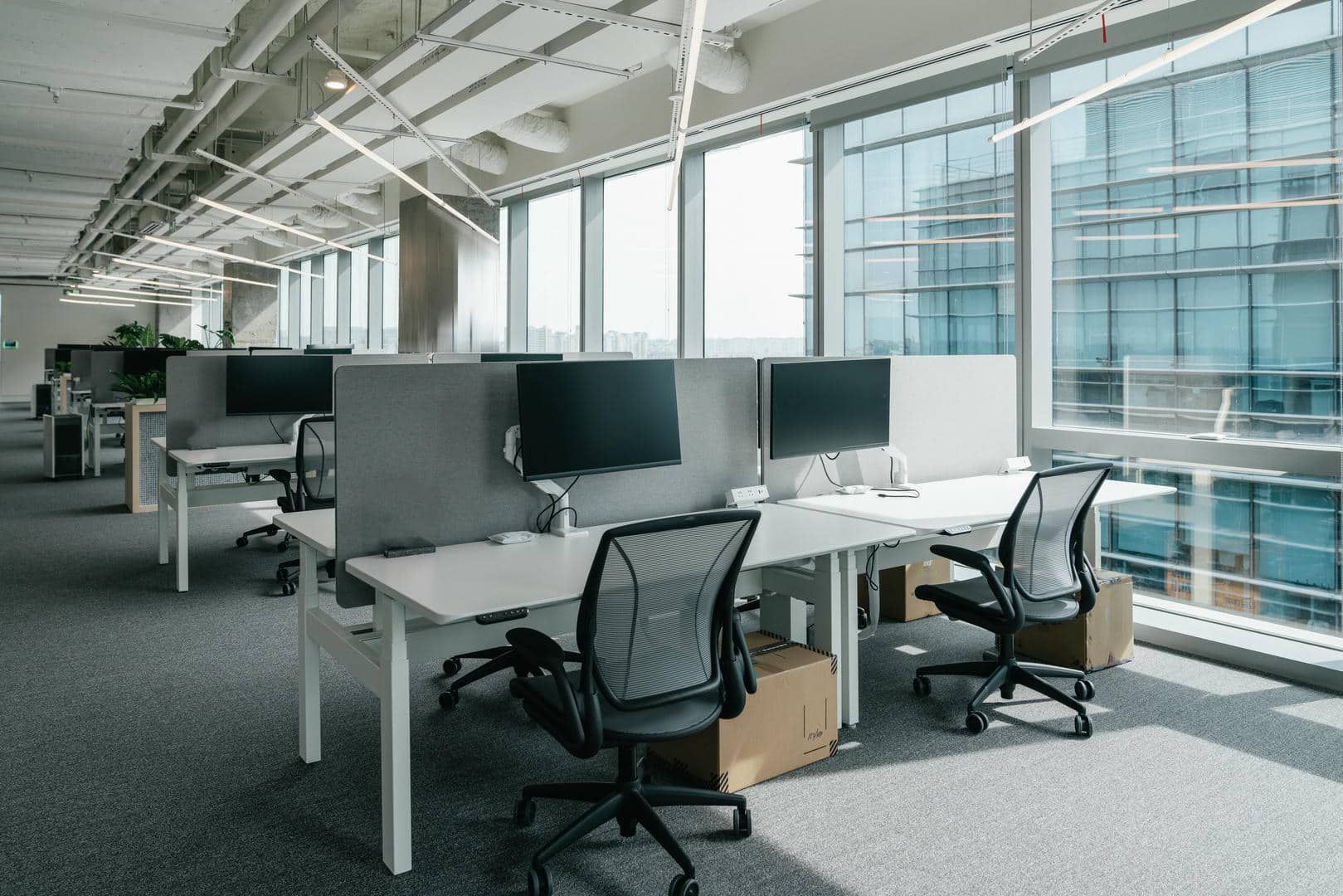 Modern office workspace with desks, chairs, computers, and large windows.