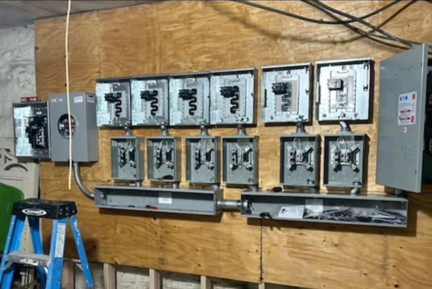 Electrical meters and breakers mounted on a plywood wall; blue ladder on left.