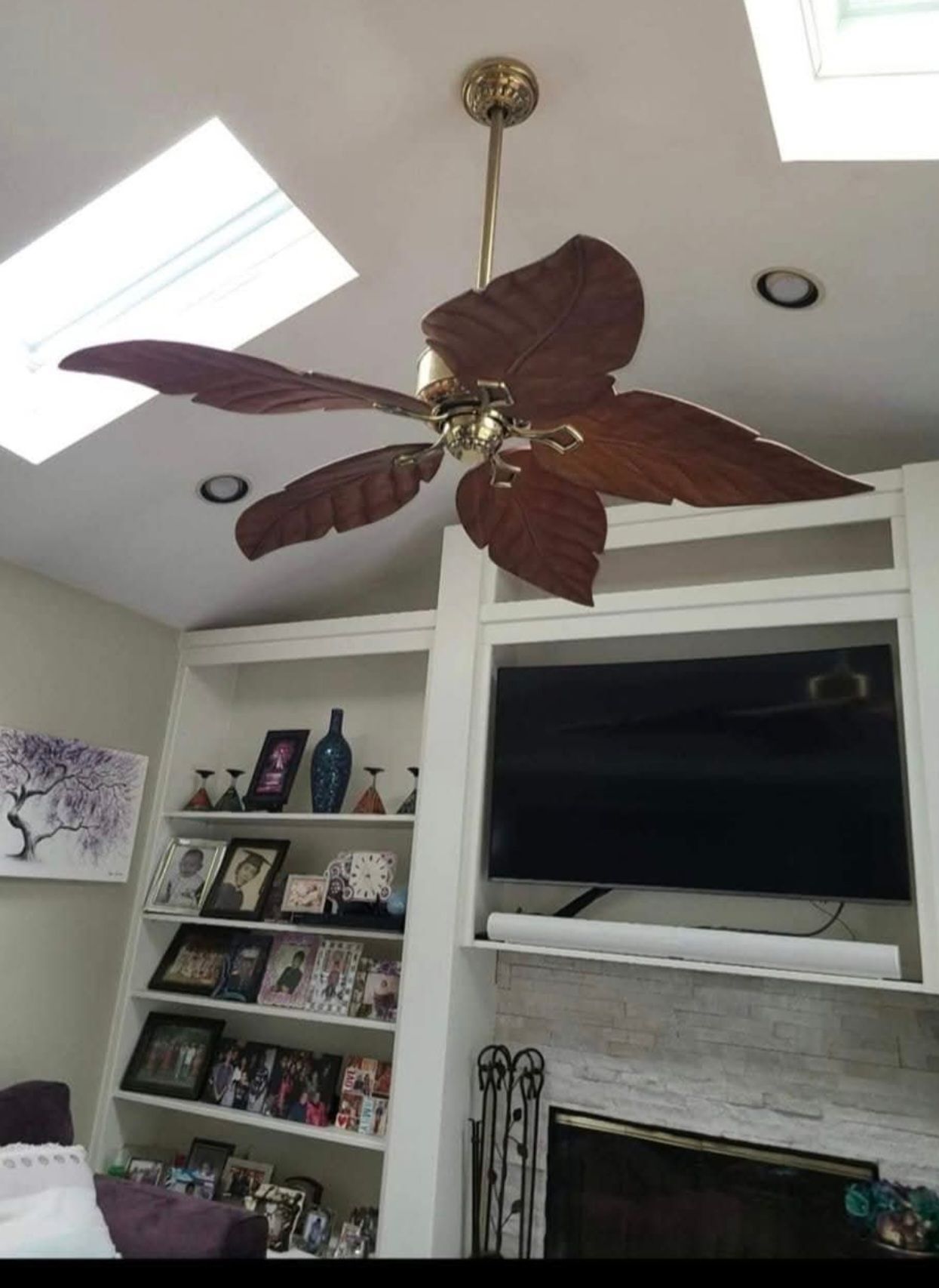 Ceiling fan with wooden blades hangs above a fireplace and shelves filled with items; sunlight streams in from a skylight.