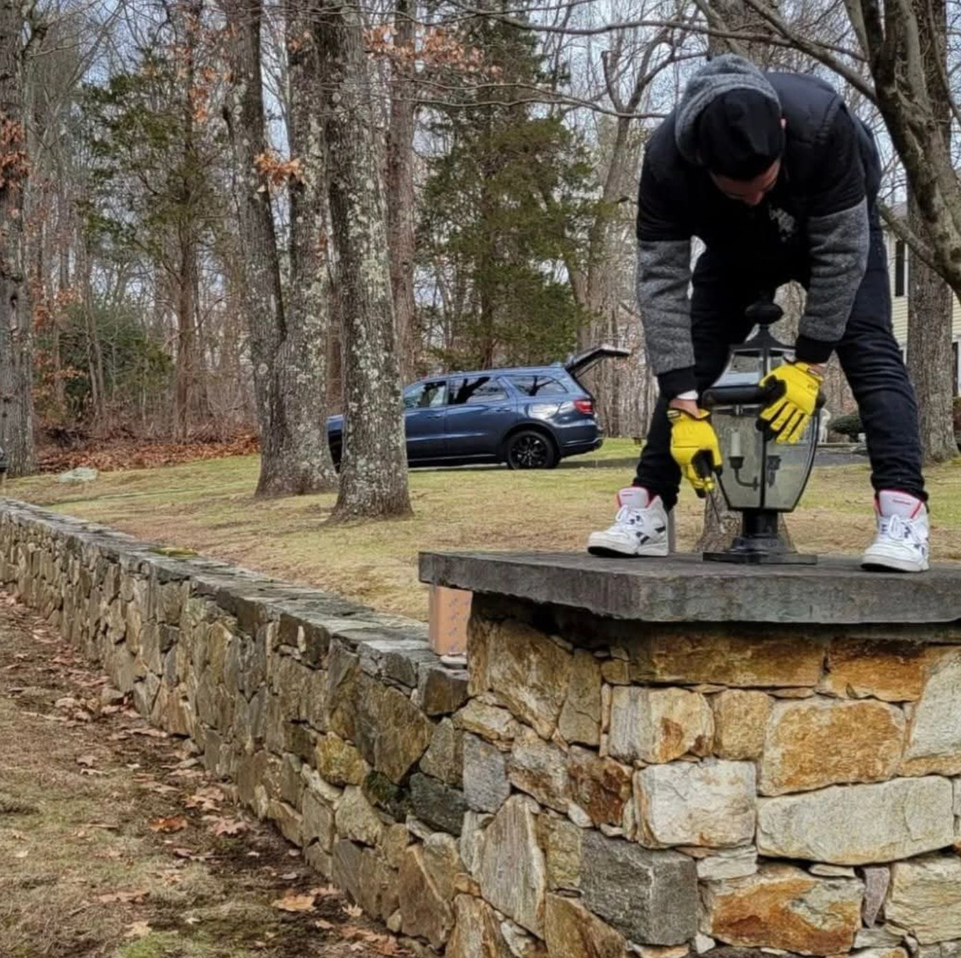 Man removing lamp from stone wall in a yard. Car and trees in the background.