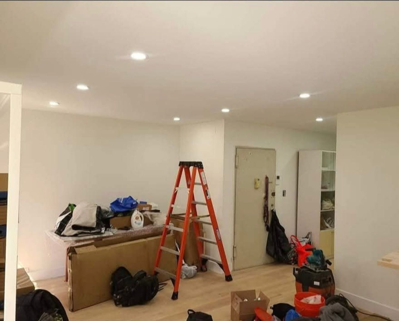 Interior room under renovation: ladder, boxes, tools, recessed lighting, white walls and ceiling, light wood floor.