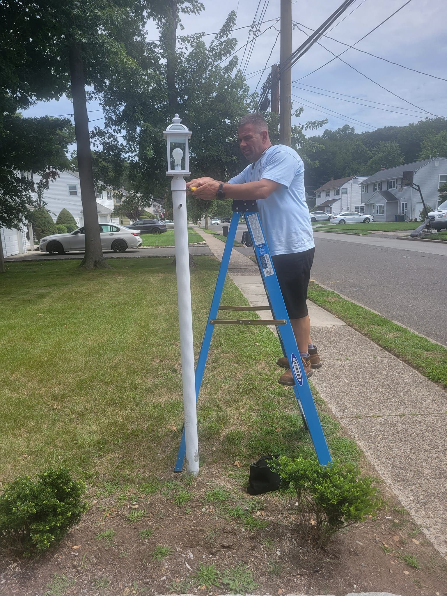 Man on ladder fixing white lamp post on a lawn. Blue ladder, green grass, suburban street.