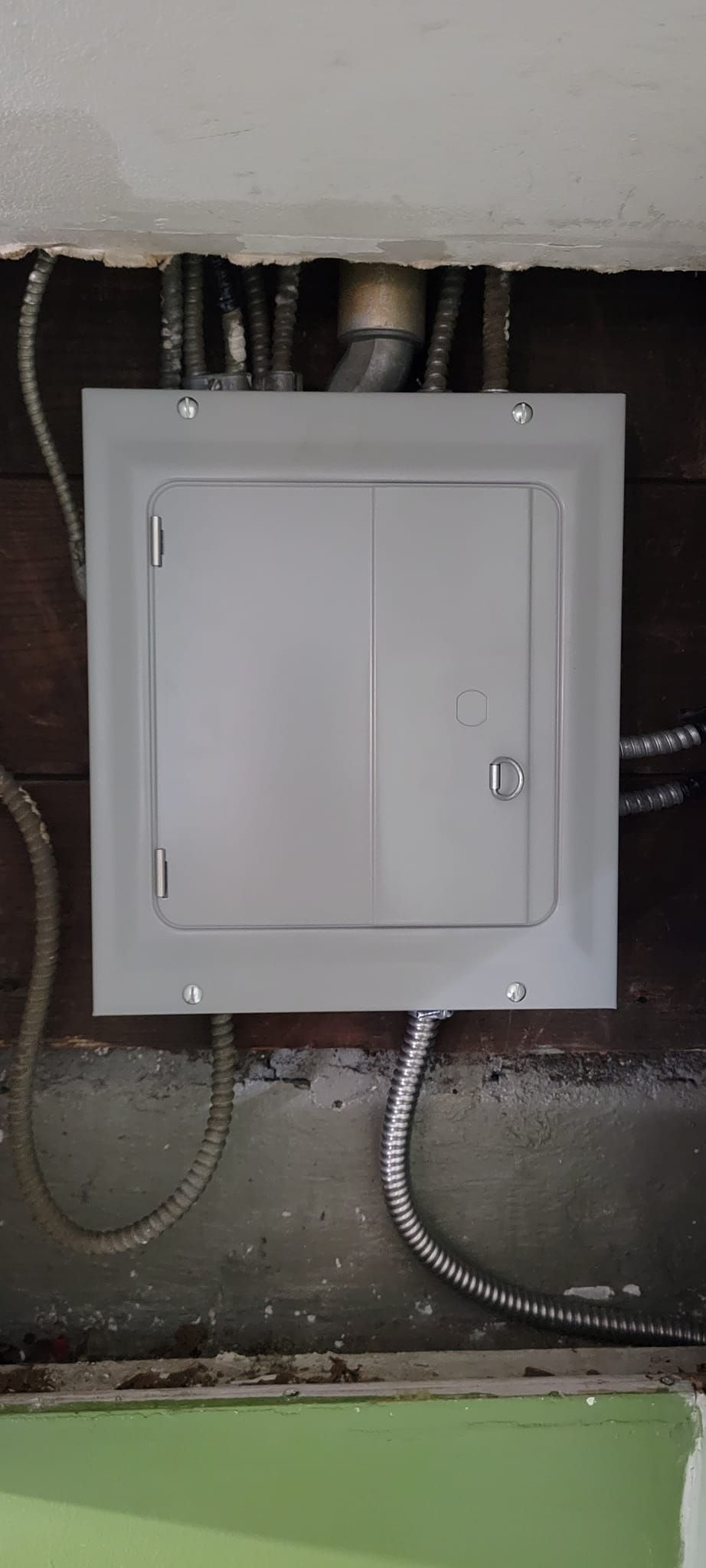 Electrical panel mounted on a dark wooden wall. Metal conduit pipes enter from above and below.