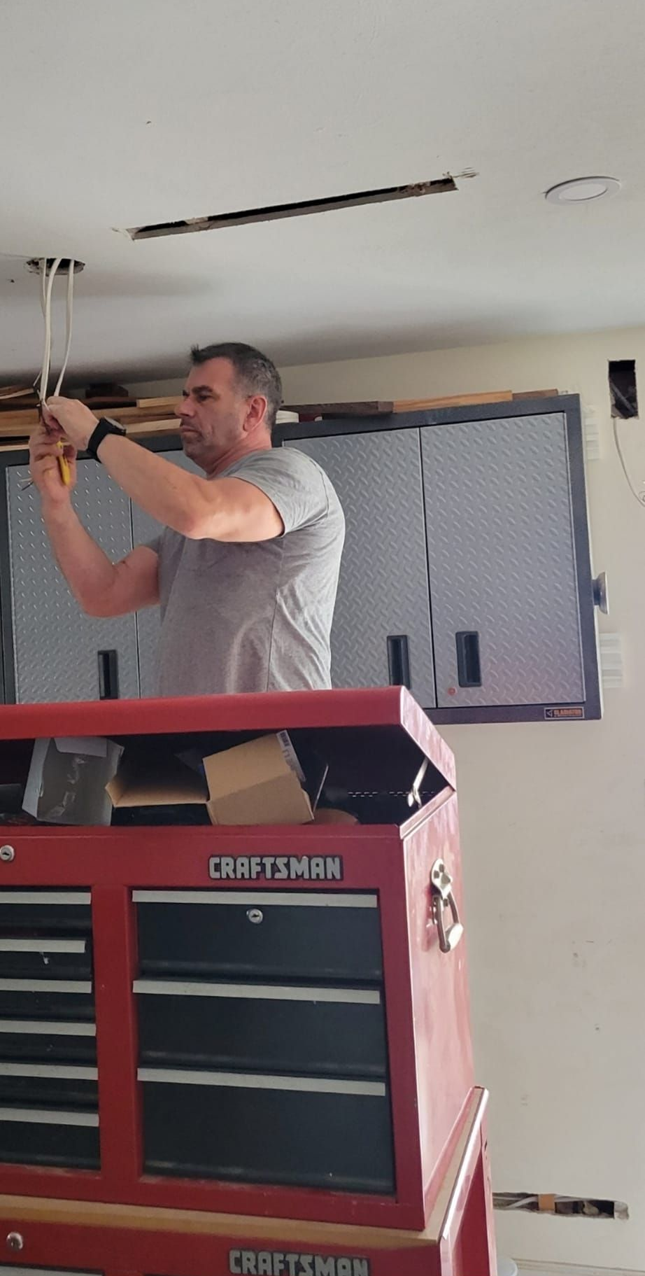 Man using tools to work on wires, standing on a red Craftsman toolbox in a room with exposed ceiling.