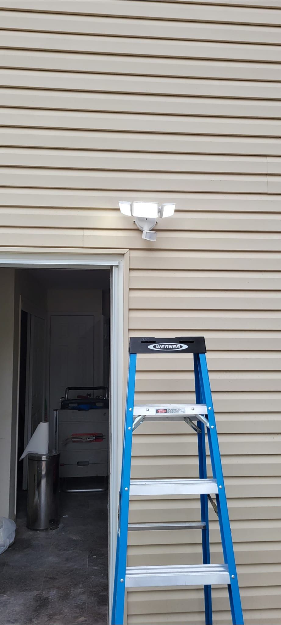 A blue ladder against a tan-sided building, with a light fixture above the doorway.