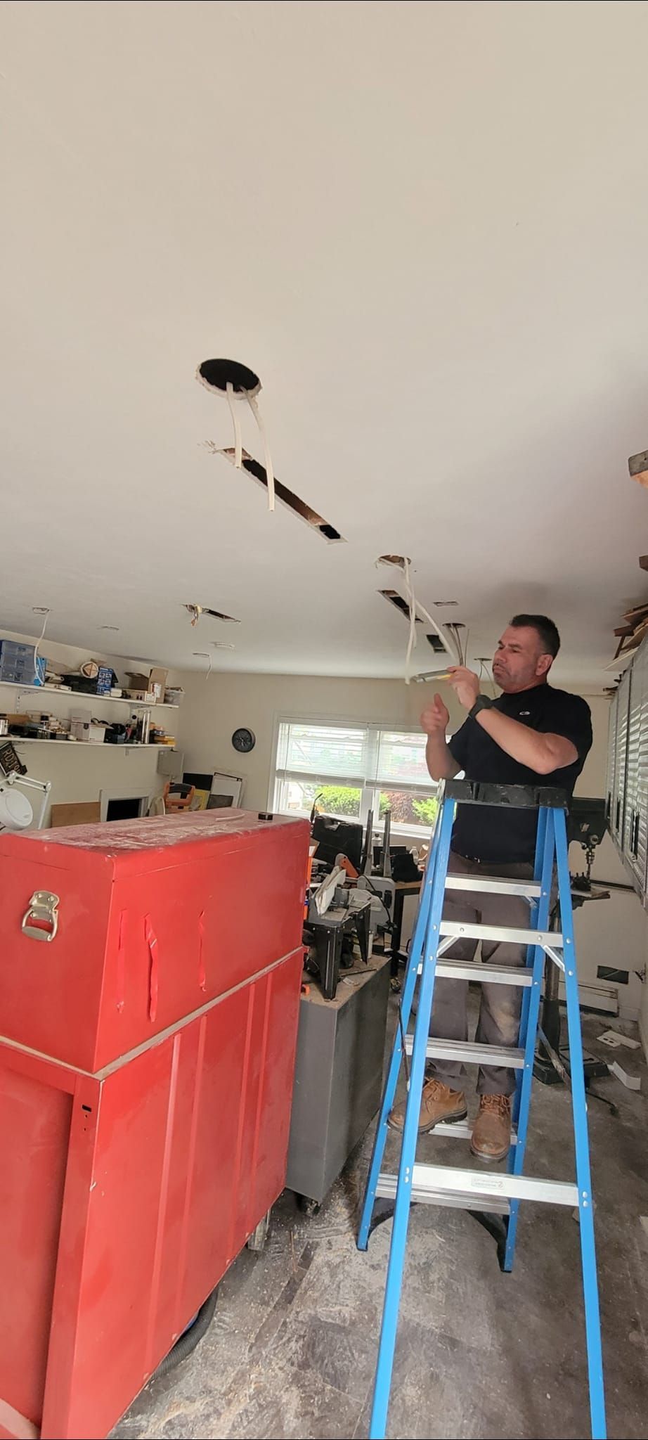 A man on a ladder repairs ceiling. He wears black shirt. Red toolbox is in the foreground.